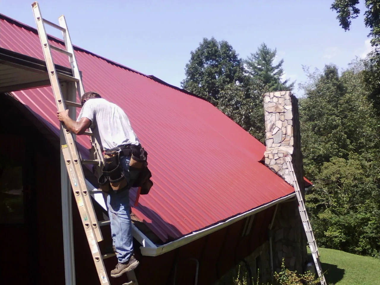 Roofer on a ladder beside a red metal roof and stone chimney — Soffit & Fascia Repair by Roofing Brothers in Welches
